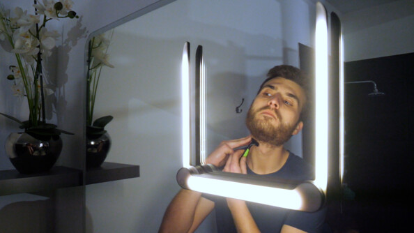 Homme se rasant de près la barbe au rasoir devant le miroir d'une salle de bains moderne sur lequel est collé la lampe LED réglable à trois bras ajustables en forme de U