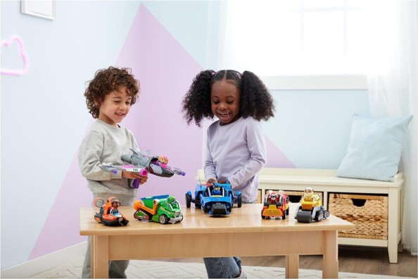 Collection des cinq véhicules Deluxe Pat' Patrouille Chase, Rocky, Marcus, Ruben, Zuma et Stella posée sur une table en bois devant deux jeunes enfants dans une salle de jeu blanche et rose lumineuse