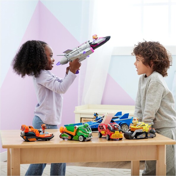 Collection des cinq véhicules Deluxe Pat' Patrouille Chase, Rocky, Marcus, Ruben, Zuma et Stella posée sur une table en bois devant deux jeunes enfants dans une salle de jeu blanche et rose lumineuse