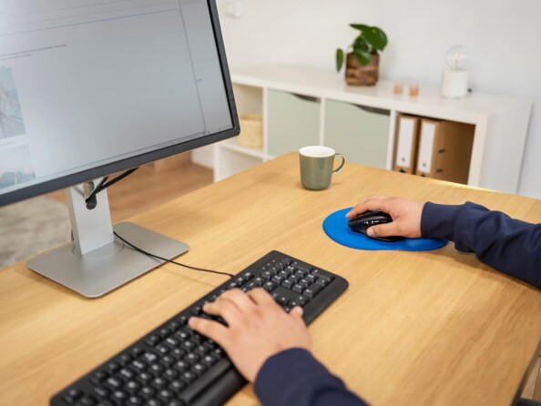 Mise en situation de l'utilisation du pad de souris bleu trust Bigfoot par un homme assis devant un bureau en bois clair avec souris sans fil dans sa main posé sur le repose-poignet du produit et main gauche tapant sur un clavie rfilaire devant un écran